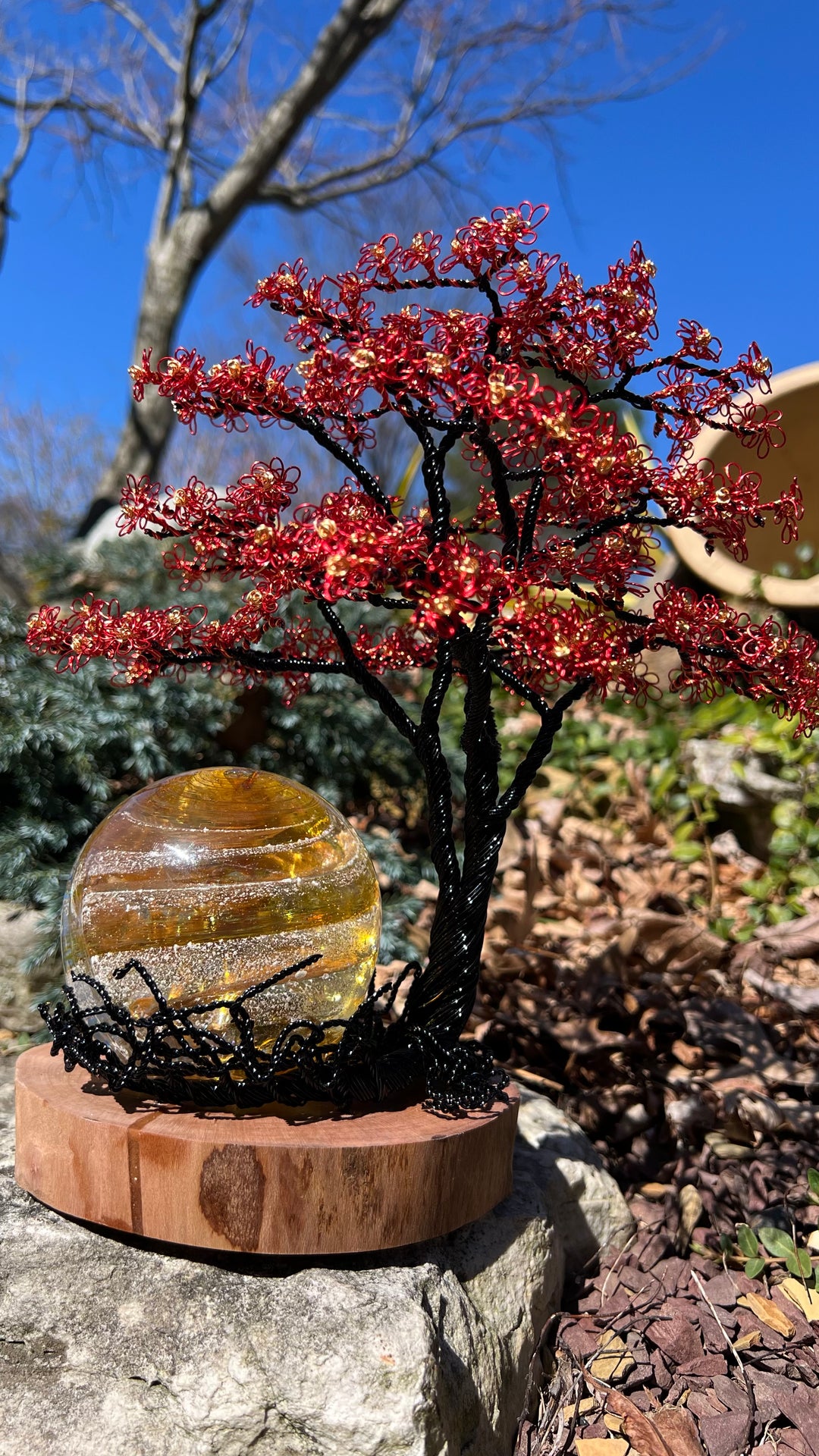 Flamboyant Tree of Life sculpture with red wire blossoms on a black trunk and a glass orb on a wooden base outdoors