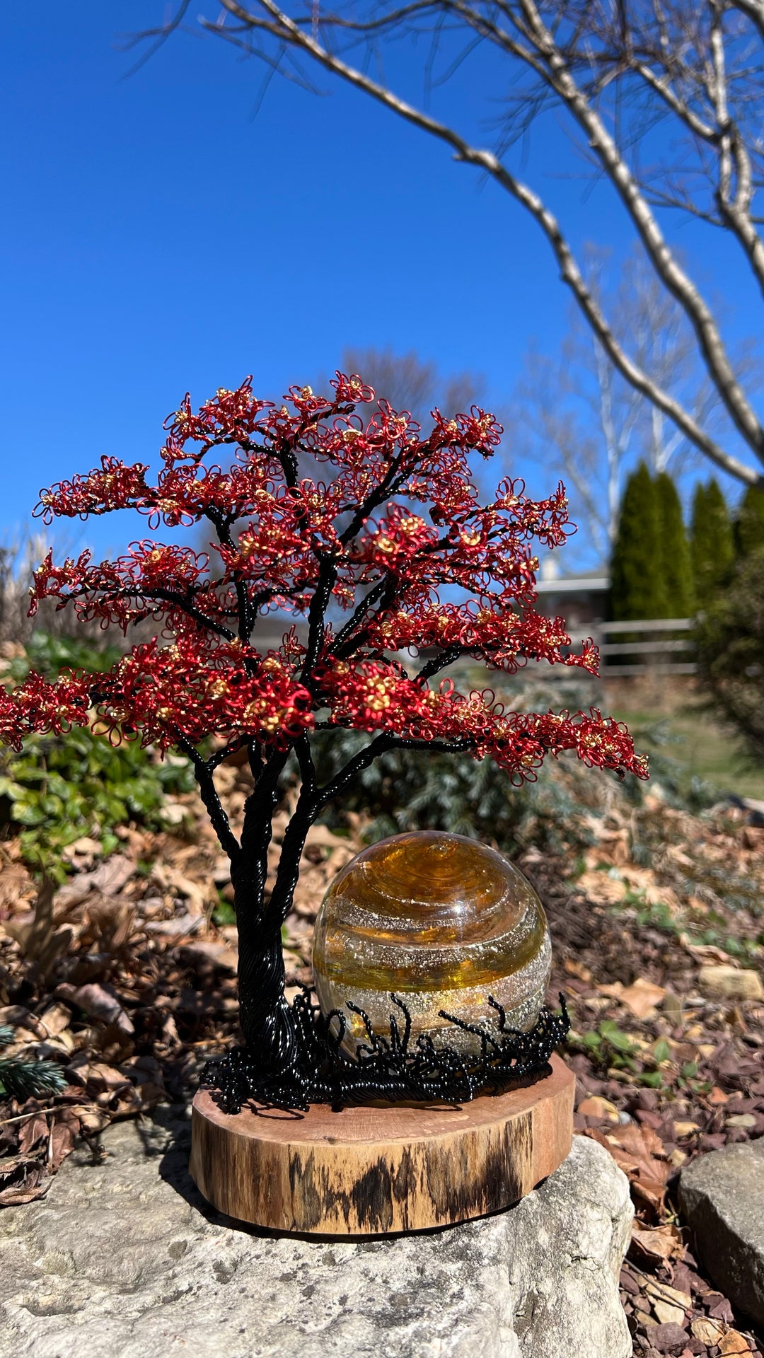 Handcrafted black wire tree with red and gold blossoms beside a 3 inch Sparkle Orb on a wooden base in a garden setting.