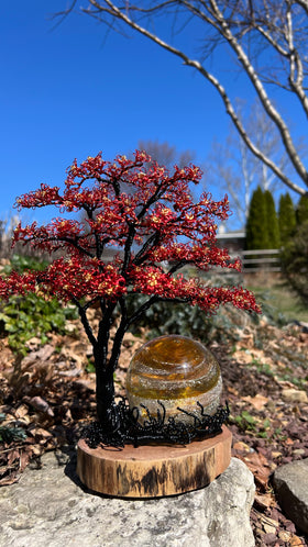 Handcrafted black wire tree with red and gold blossoms beside a 3 inch Sparkle Orb on a wooden base in a garden setting.
