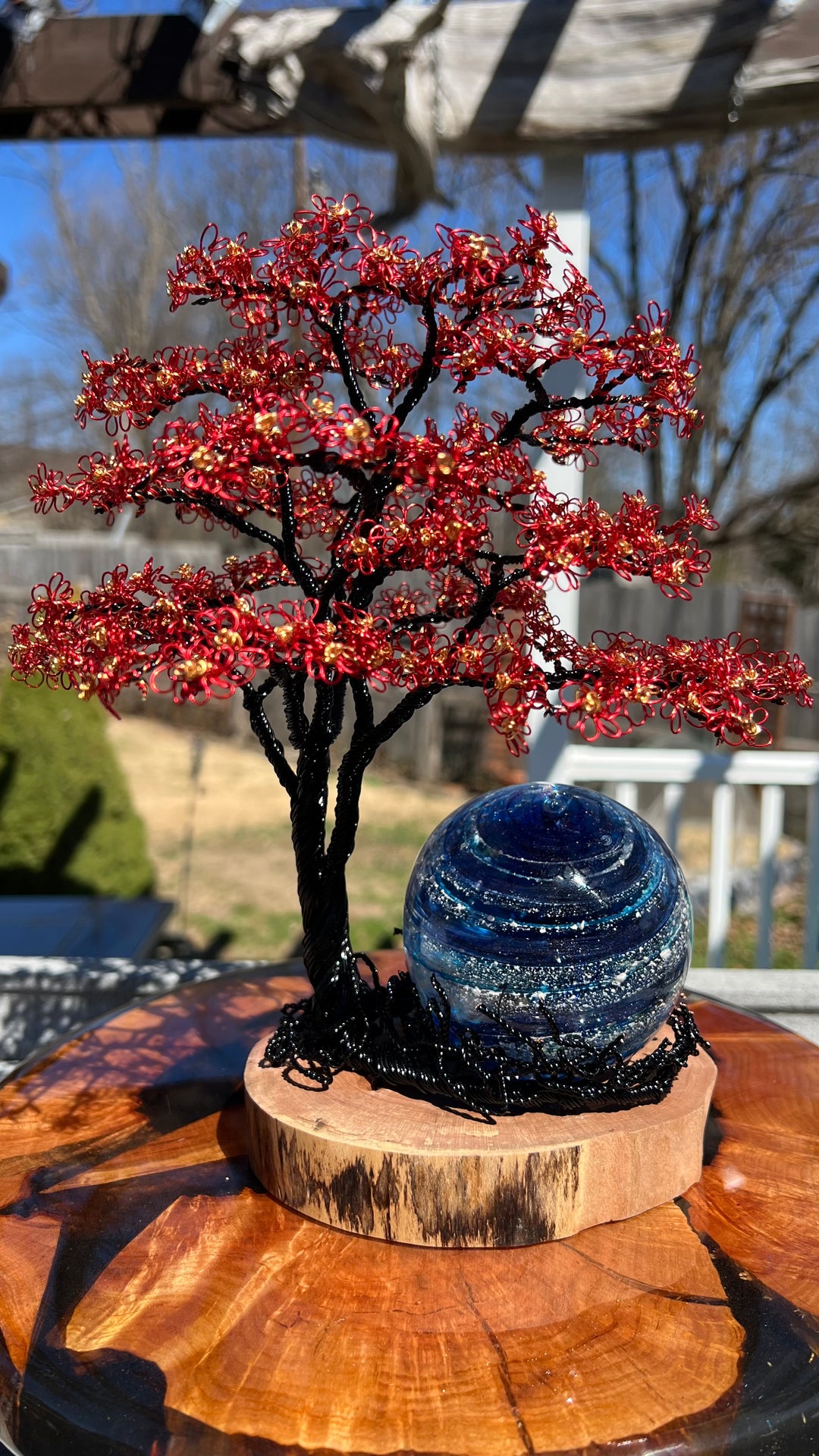 Flamboyant Tree of Life wire sculpture with red blossoms and a blue Sparkle Orb on a round wooden base outdoors