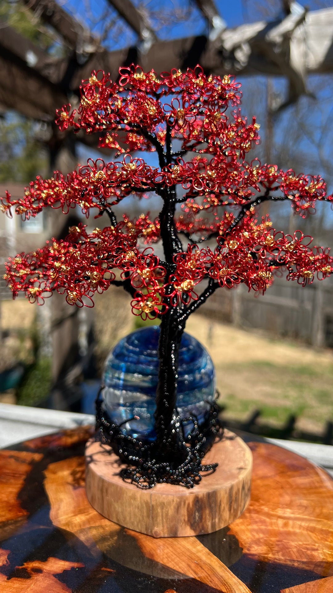 Flamboyant Tree of Life wire sculpture with red flower clusters and gold centers on a black trunk, set on a wooden base beside a blue Sparkle Orb.