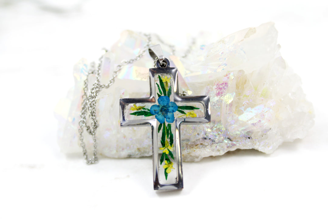 Crystal resin cross pendant with a blue flower center and green leaves inside a silver frame on a silver chain, set against a white crystal cluster.