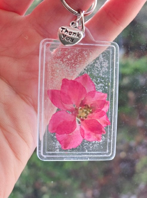 Clear plastic framed keychain with a pink dried flower and cremation ash specks, plus a Thank You charm and ring.