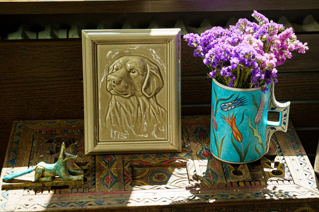 Ceramic Labrador frame urn with embossed dog portrait leans on a wood surface beside a turquoise vase with purple flowers.