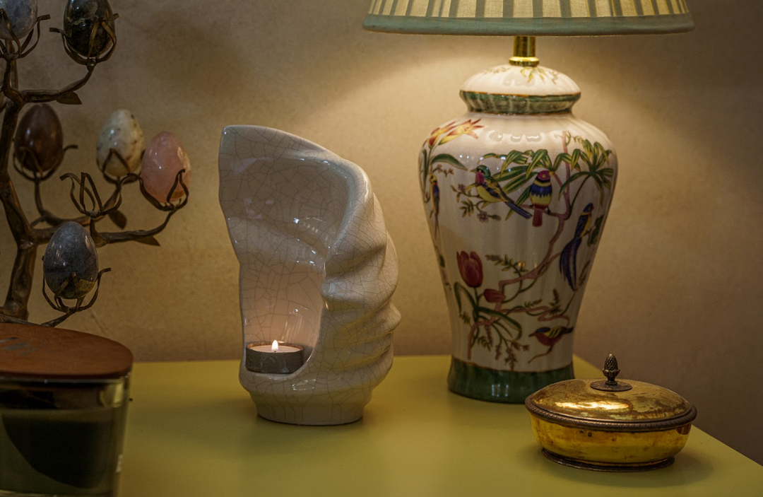 White crackled ceramic candle urn with inner cavity holding a small tealight on a yellow table beside a decorative vase.