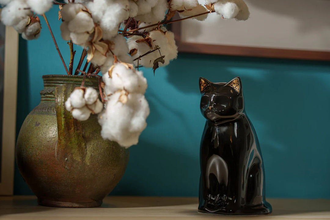 Glossy black Meow cat ceramic urn displayed on a shelf beside a ceramic vase with cotton stems