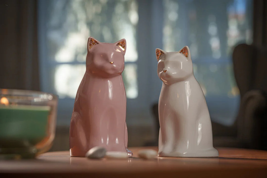 Pair of Meow cat ceramic urns in pink and white on a wooden table with a candle holder in the background.