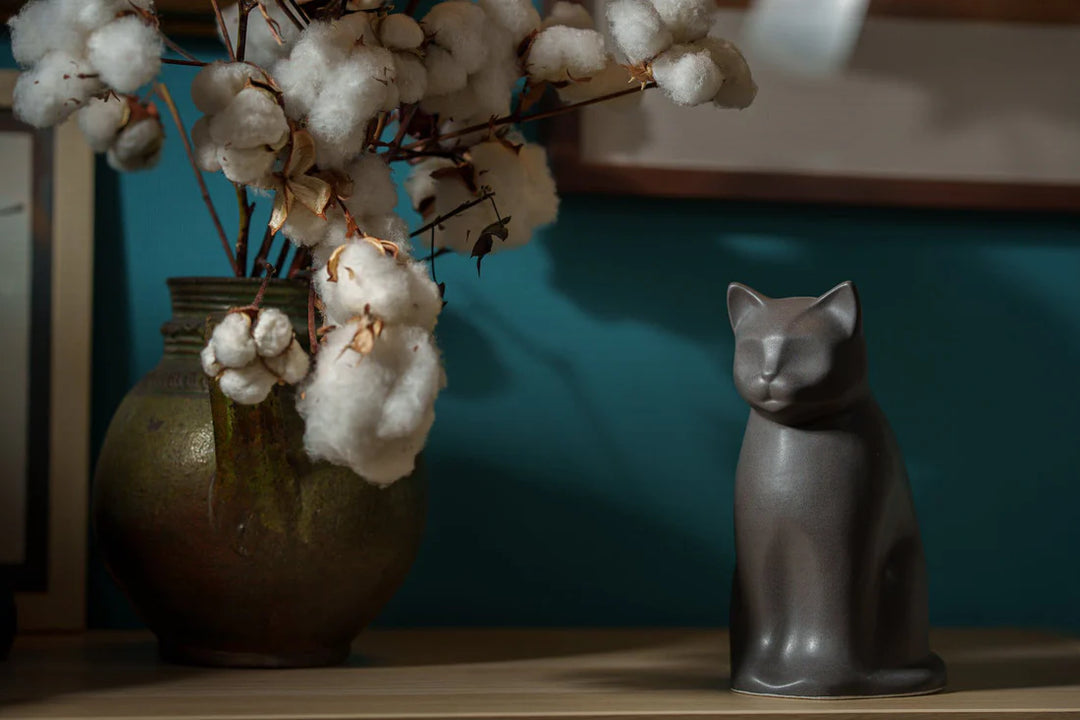 Gray cat shaped ceramic urn on a shelf beside a rustic vase with cotton stems.