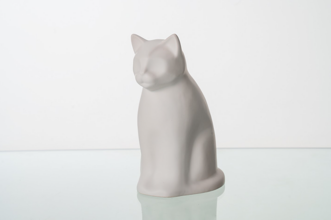 White ceramic cat urn sitting on a glass surface against a white background.