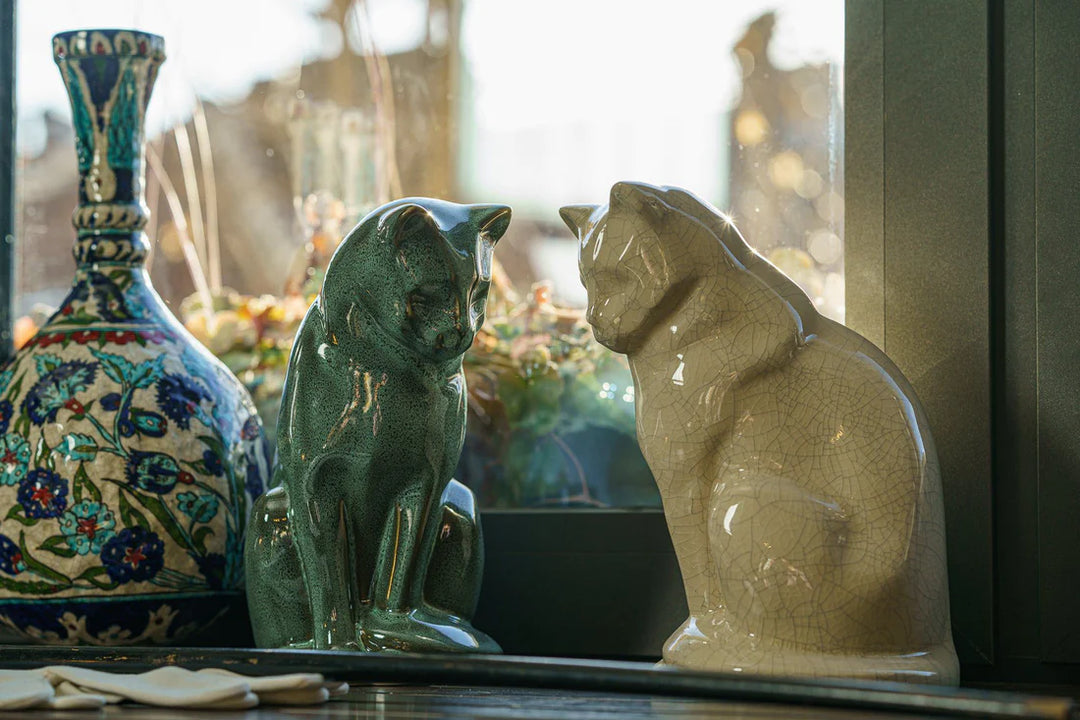 Two cat ceramic urns in green and beige finishes sit together on a shelf beside a decorative patterned vase.