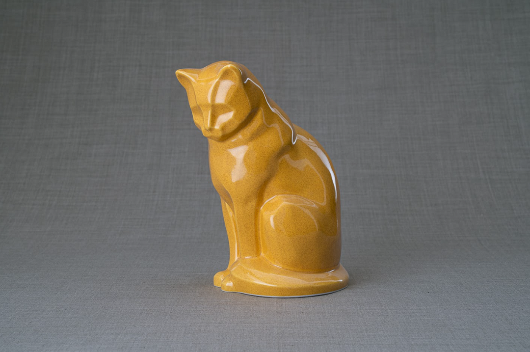 Mustard yellow ceramic cat urn in a seated pose on a neutral fabric backdrop.