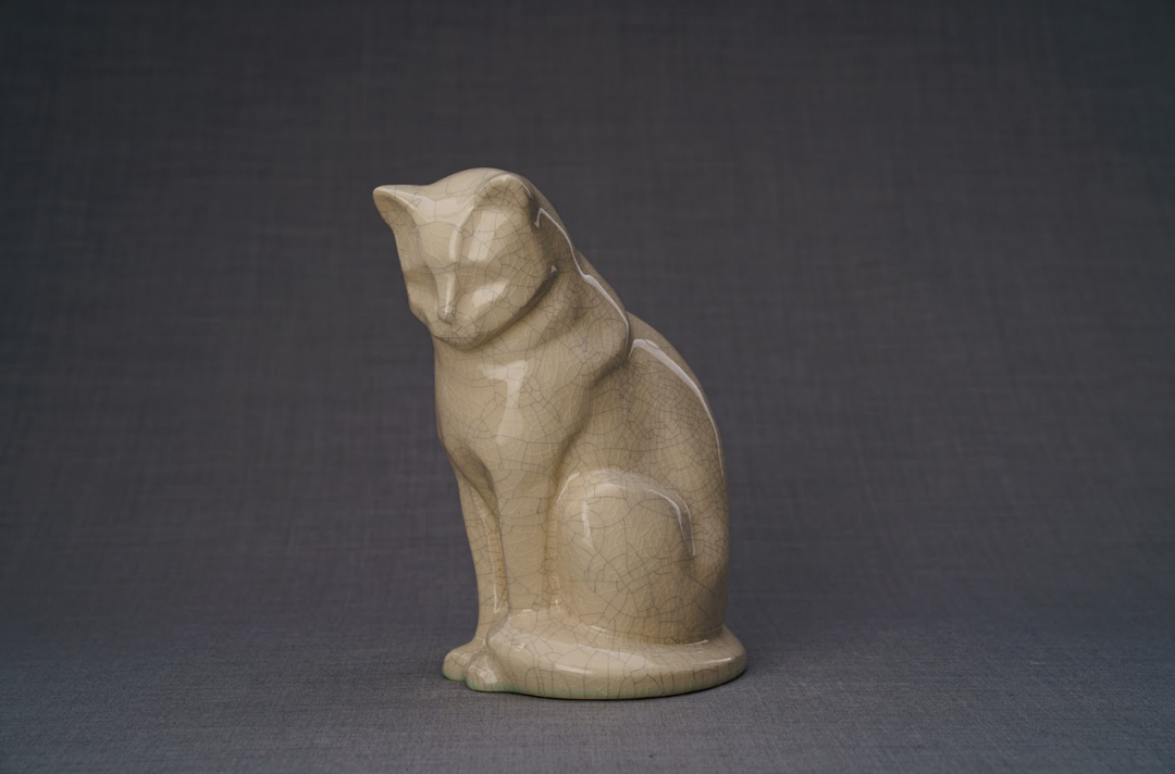 Beige ceramic cat urn in a seated pose with a glossy crackled glaze against a dark backdrop.