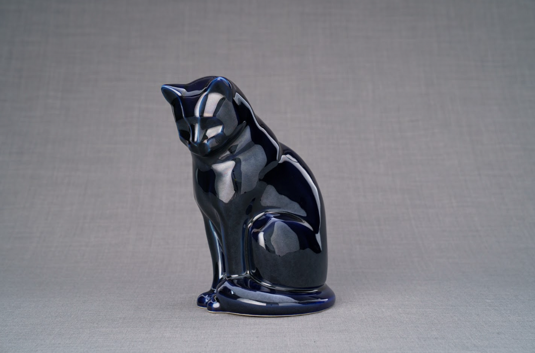 Dark blue glossy ceramic cat urn in a seated pose on a gray studio backdrop.