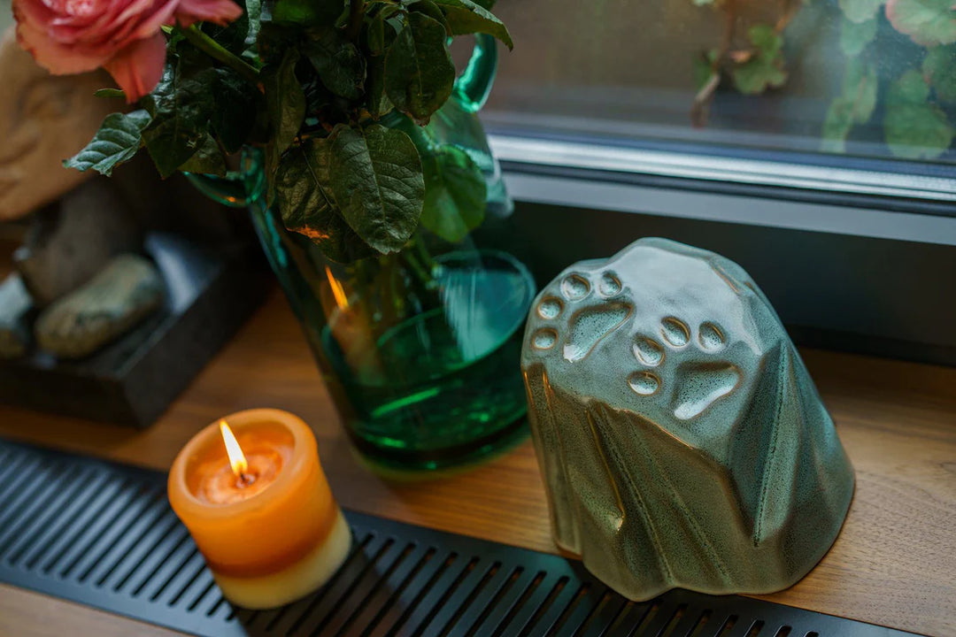Green glaze paw print ceramic urn with a stonelike shape sits on a windowsill beside a candle.