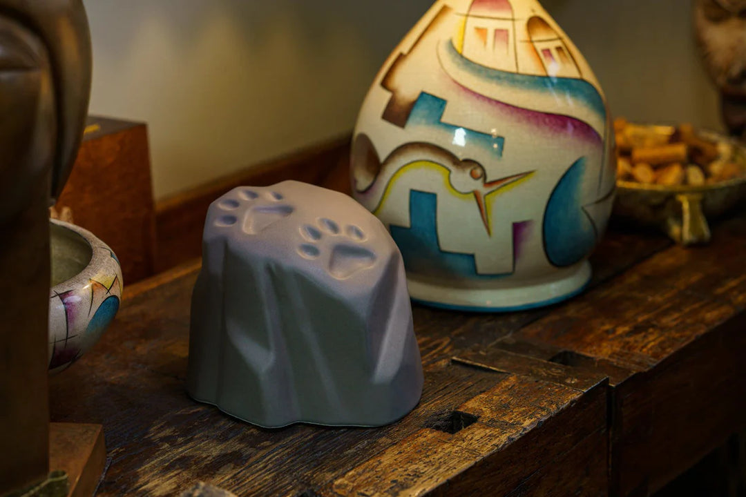 Stone shaped paw print ceramic urn in matte gray sits on a wooden surface with colorful pottery in the background
