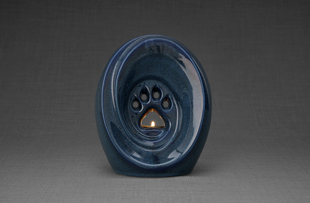Blue glazed ceramic paw print pet urn with a candle holder and a lit candle against a neutral gray backdrop