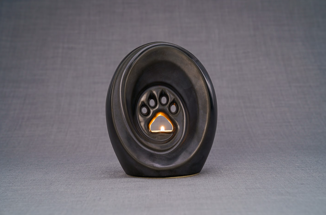 Dark ceramic paw print pet urn with built in candle holder and lit flame on a gray surface
