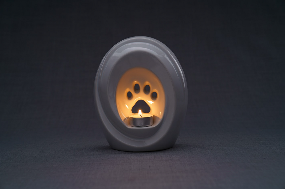 Light gray ceramic paw print pet urn with built in candle holder and a lit tealight against a dark background