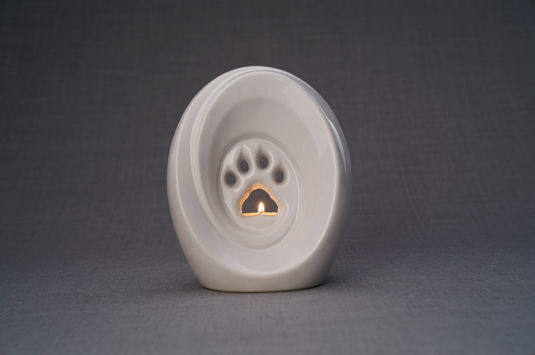 White paw print ceramic pet memorial urn with built in candle holder and a small flame visible inside.