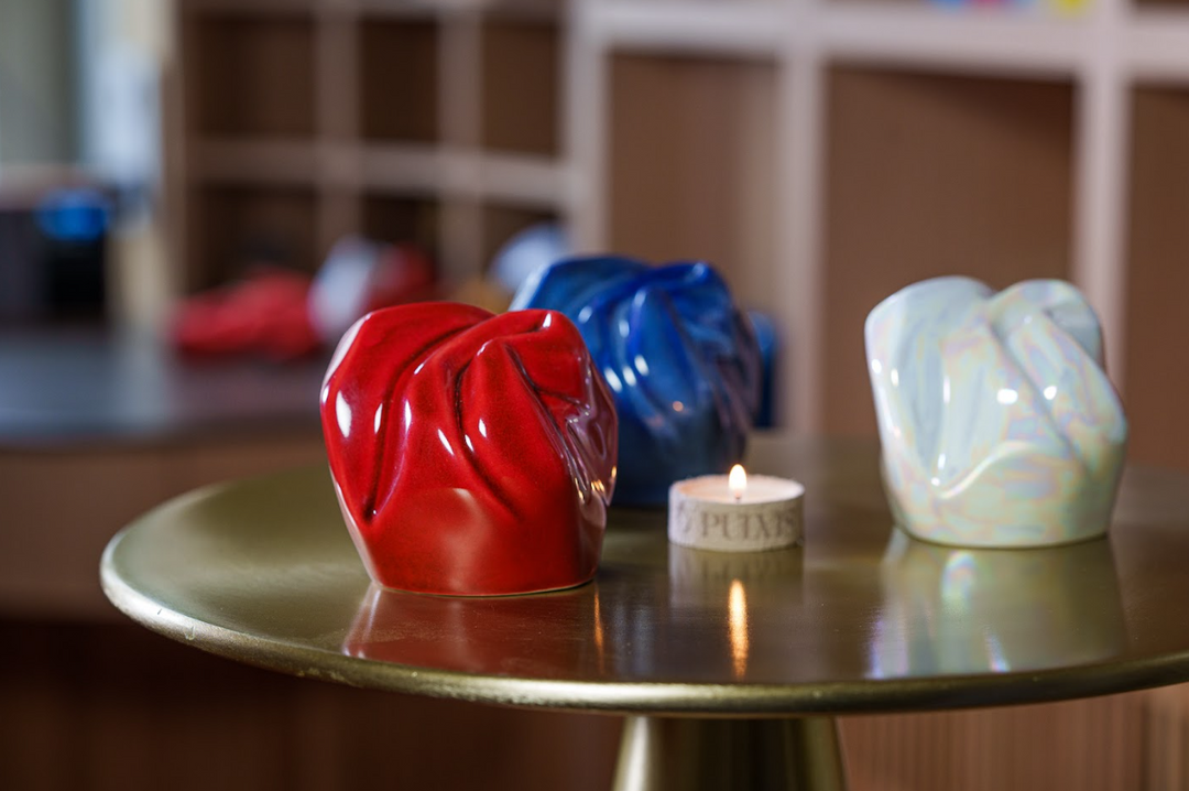 Red blue and pearlescent white ceramic keepsake urns on a round brass table with a small candle.