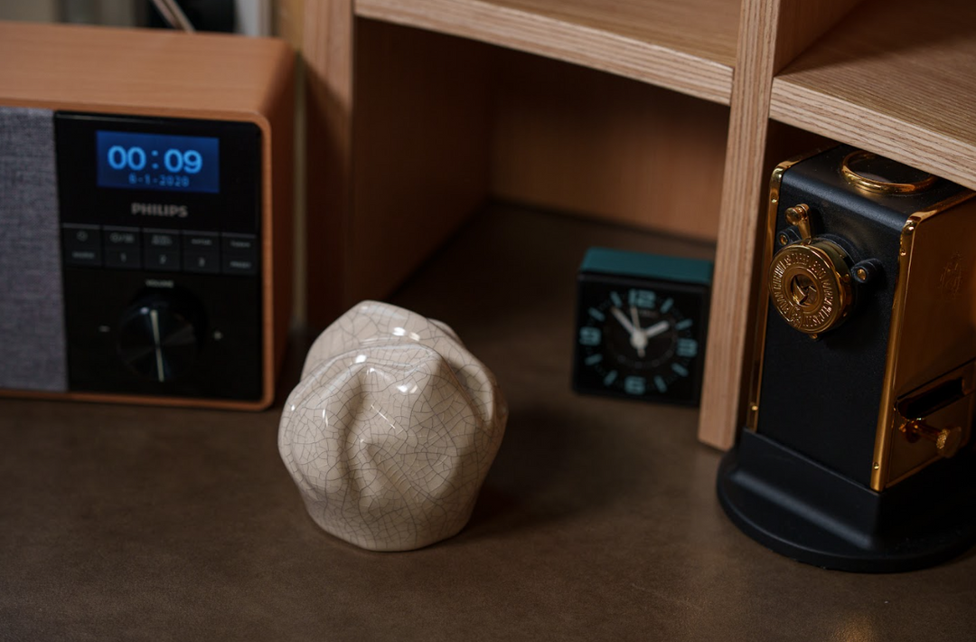 Crackled pale pink ceramic keepsake urn on a brown desk beside a vintage camera and small clock.
