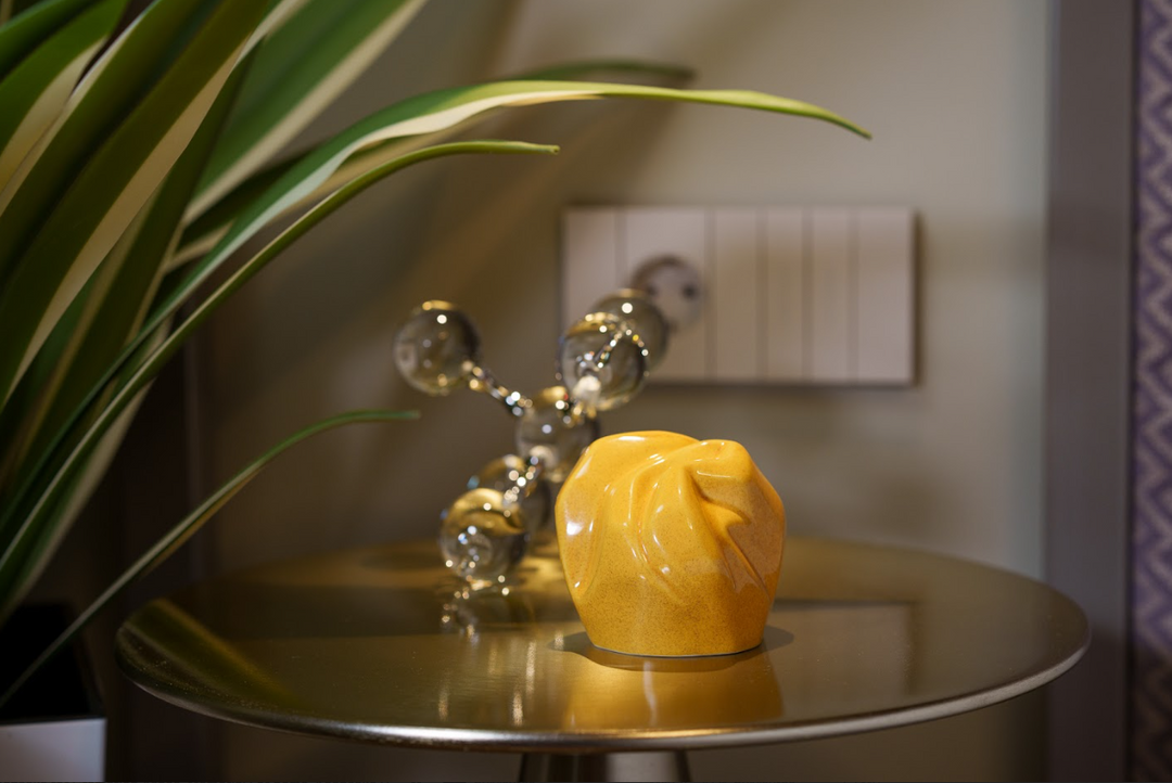 Precious ceramic keepsake urn in mustard yellow resting on a round table with a plant and decorative glass sculpture