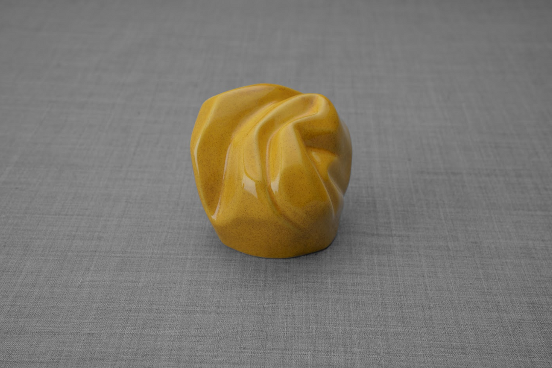 Yellow ceramic keepsake urn with flowing folded design on a gray surface