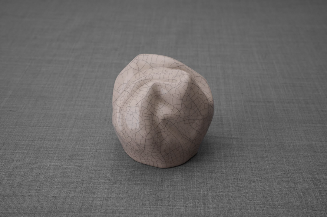 Crackled pale pink ceramic keepsake urn placed on gray fabric surface.