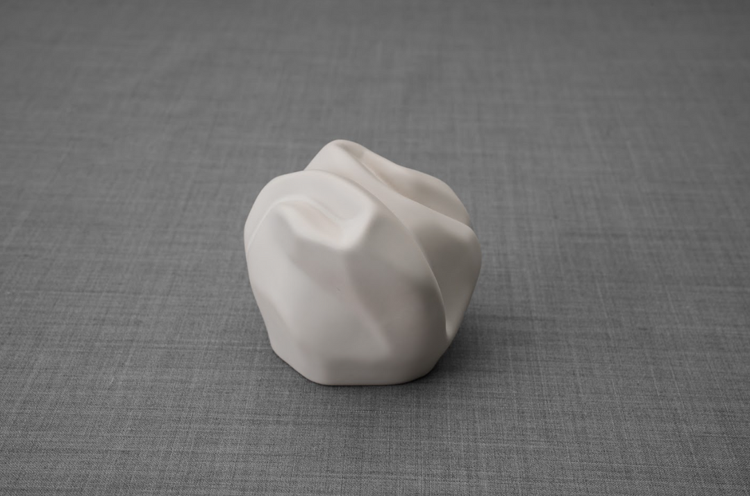White ceramic keepsake urn with a sculpted folded design shown on a gray fabric background