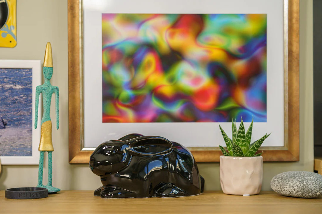 Glossy black rabbit shaped ceramic urn on a wooden surface with a colorful abstract framed artwork behind.
