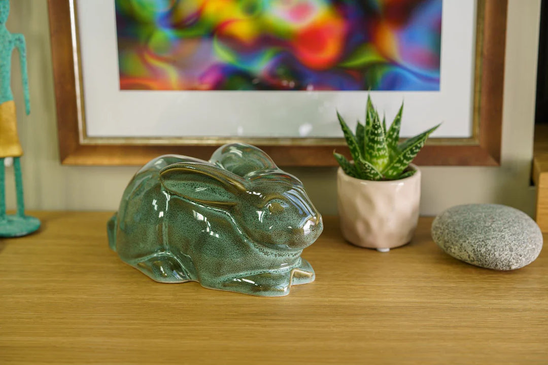 Rabbit ceramic urn with glossy green glaze sits on a wooden surface beside a small potted plant and a smooth stone.