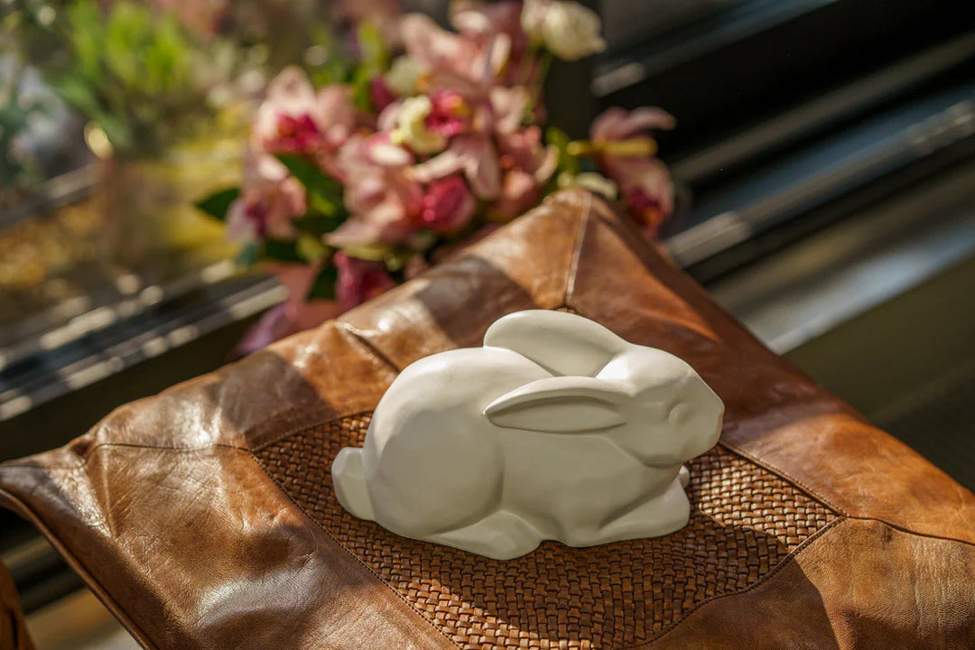 Glossy black rabbit urn set on a brown textured surface with blurred flowers in the background