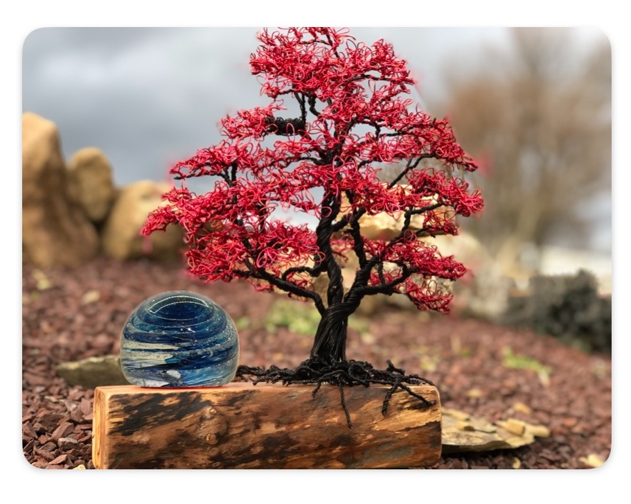 Red maple wire tree on a wooden base with a blue glass orb in the foreground outdoors.