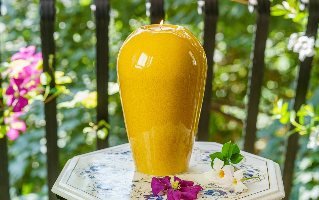Serenity yellow ceramic urn with a top tea light holder and lit candle on a floral tiled table outdoors.