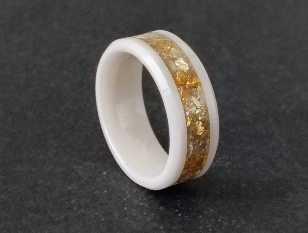 Polished cremation ring with a white band and gold ash frit encased in glass, shown on a dark surface
