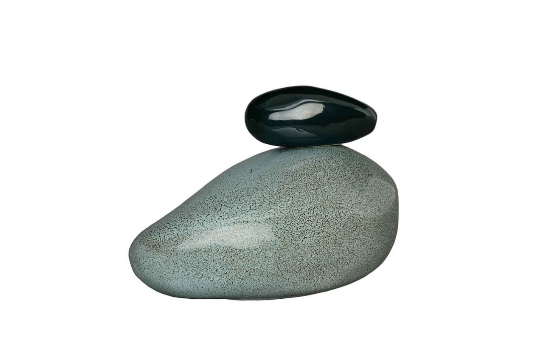 Speckled gray green teardrop shaped stone with a dark smooth rock resting on its summit against a white background