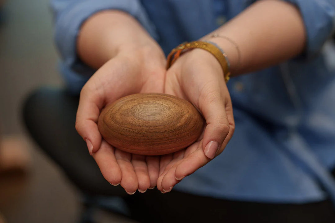 Hands cradle a smooth oval wooden keepsake urn with warm natural wood tones.