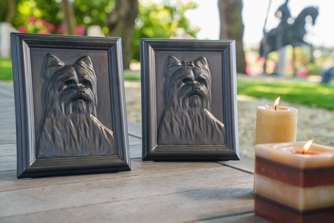 Two dark framed Yorkie relief portraits on a weathered wooden surface with candles in the foreground.