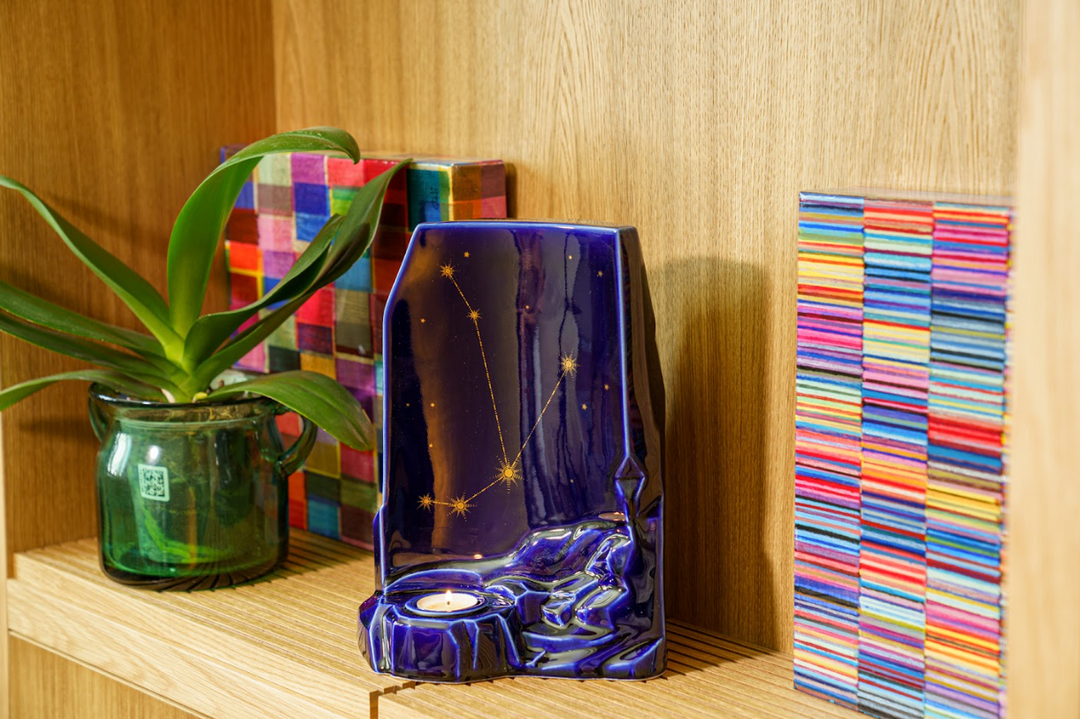 Blue celestial ceramic urn with constellation design and candle holder, placed on a wooden shelf beside a green plant.