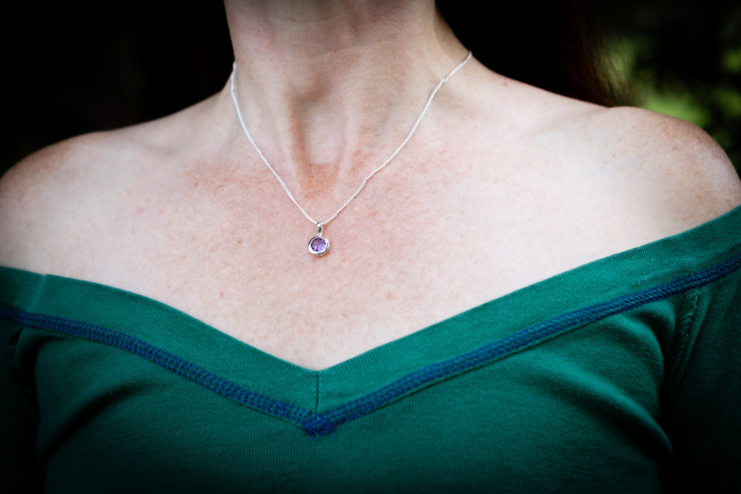 A close-up of a person's neck and shoulders wearing a delicate silver chain amethyst cremation necklace with a small round purple gemstone pendant, paired with a green off-the-shoulder top.