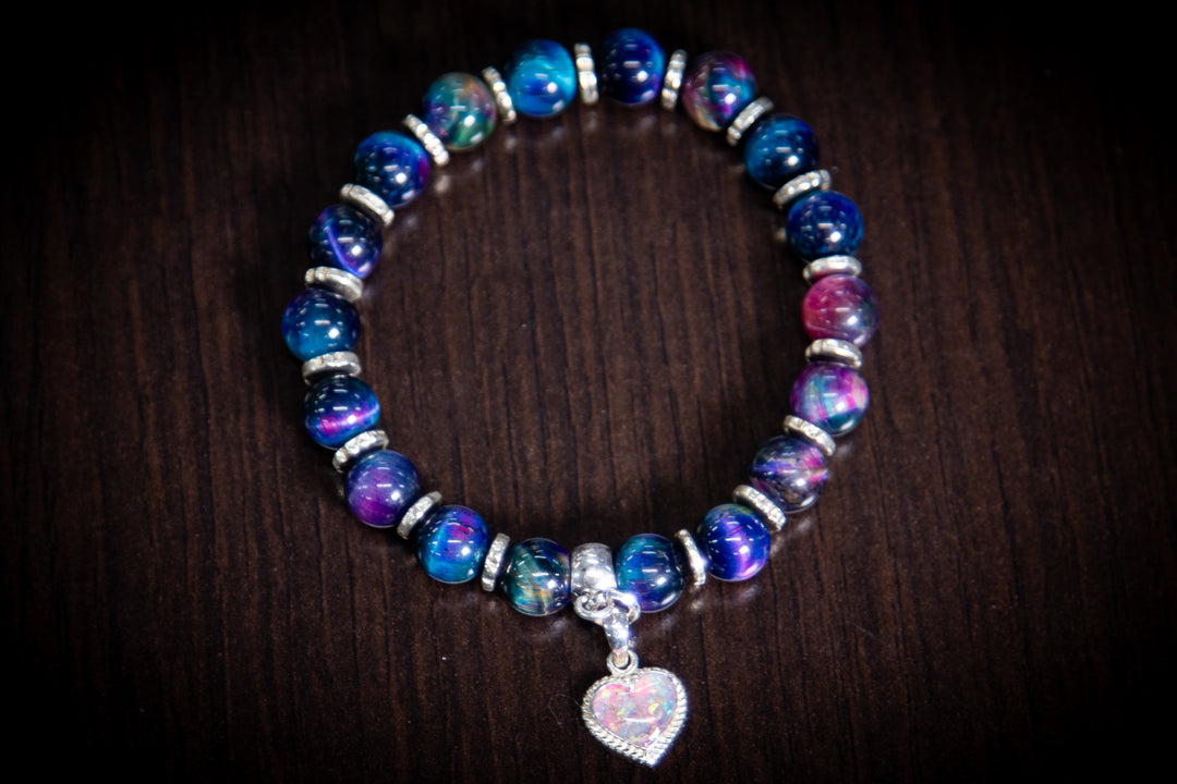 Rainbow Tiger Eye cremation bracelet with a sterling silver heart charm filled with crushed opal ashes on a dark wood surface
