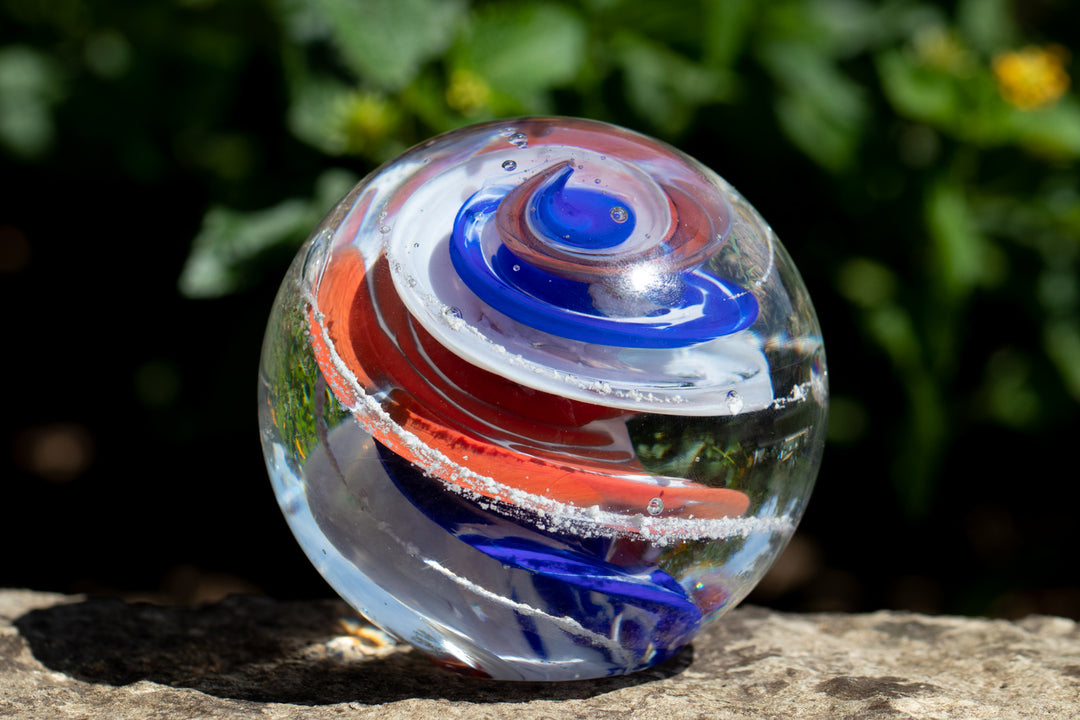 Multicolored glass cremation orb with blue and red swirls rests on a rock outdoors.