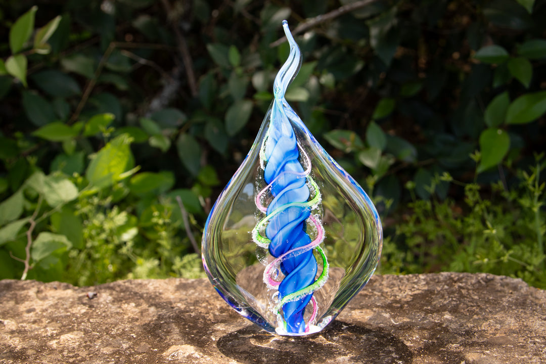 Clear glass teardrop memorial flame with two cremation ash spirals around a blue core, shown outdoors.