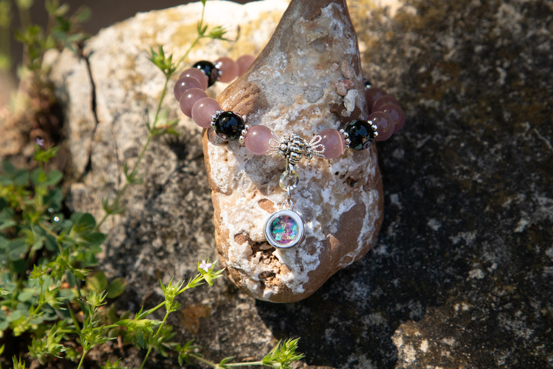Black flower and glass beaded bracelet with a silver cremation ashes charm and iridescent round pendant resting on a rock.