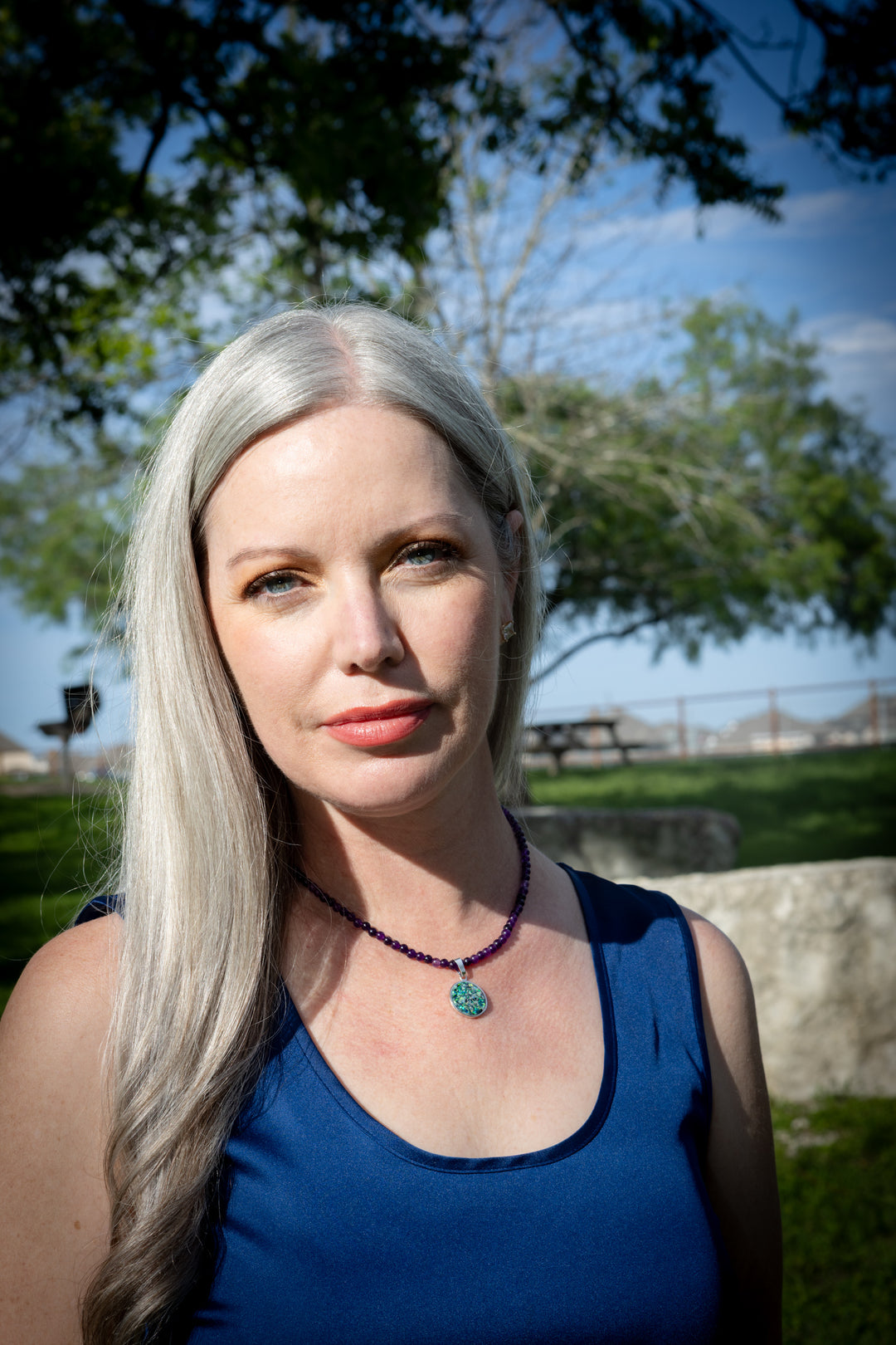 Woman wearing cremation necklace with long blonde hair wearing a blue top outdoors with trees and sky in the background