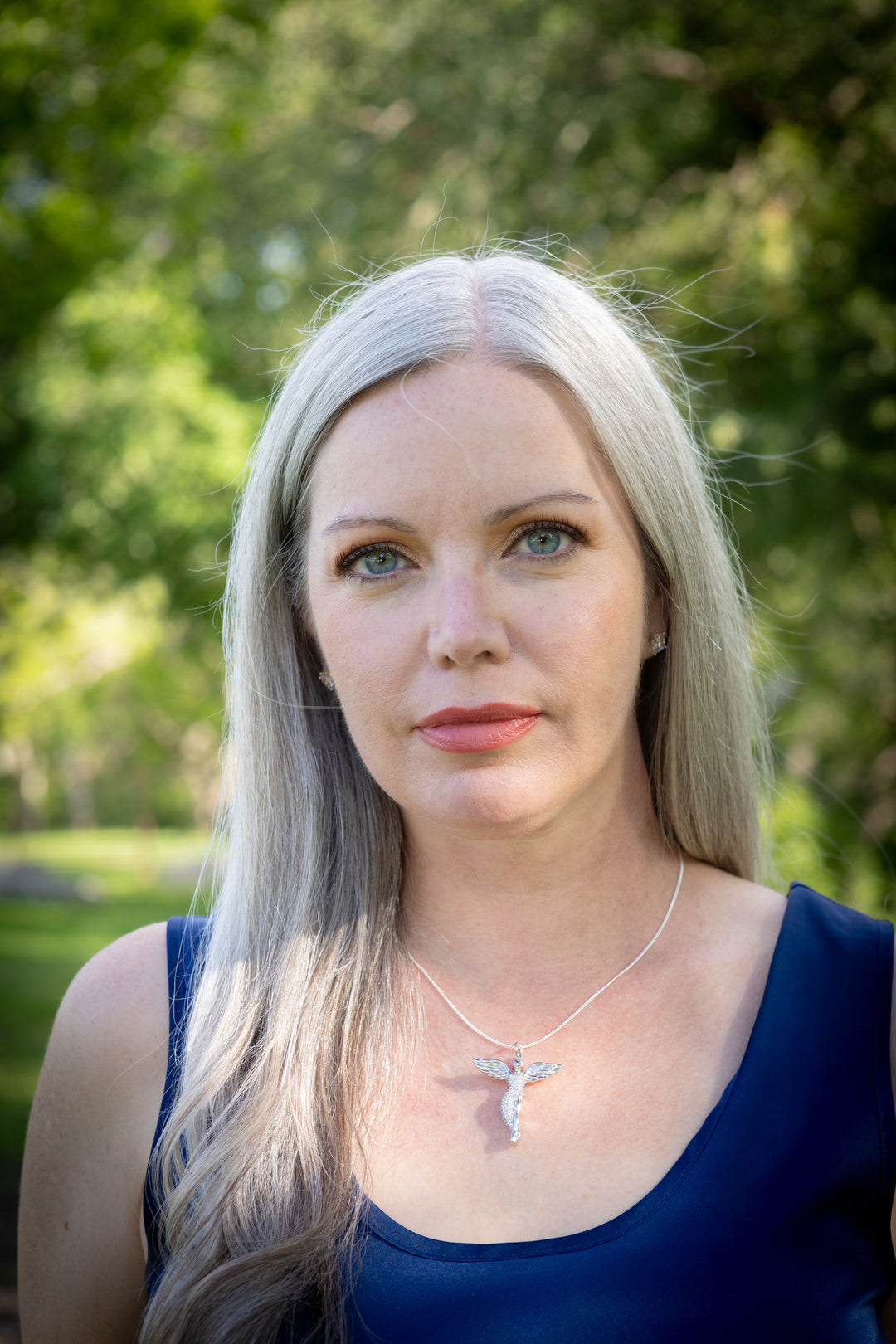 guardian angel pendant with ash worn on Woman with long blonde hair wearing a blue top and necklace in a park setting