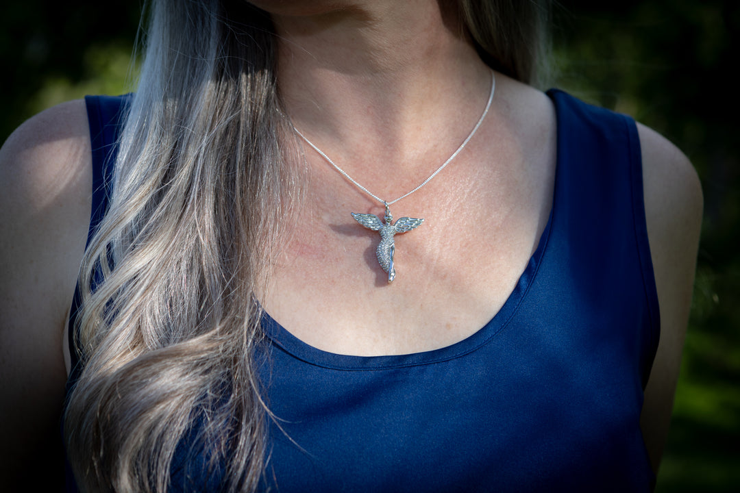 guardian angel pendant with ash worn on Woman with long blonde hair wearing a blue top and necklace in a park setting
