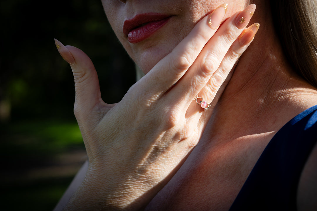 Close-up of a woman's hand wearing a filigree heart ring, covering her mouth with a blurred natural background