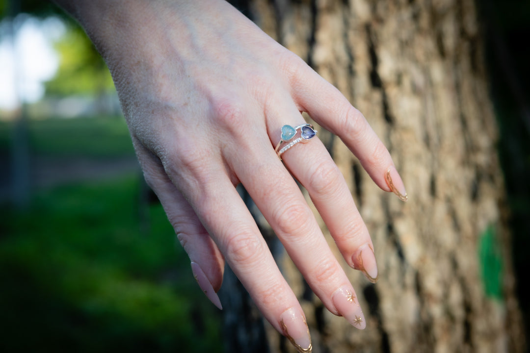 Hand wearing a double heart cremation ring with a heart and blue stone against a blurred natural background