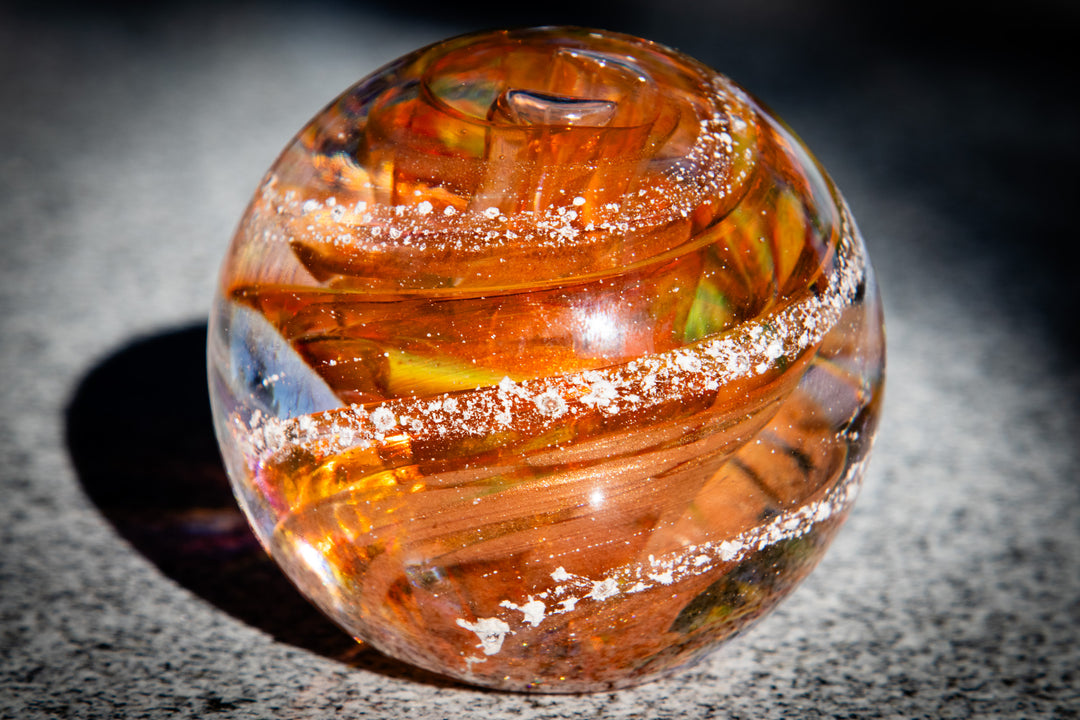 Amber orange swirl glass paperweight with ash inclusions on a speckled stone surface